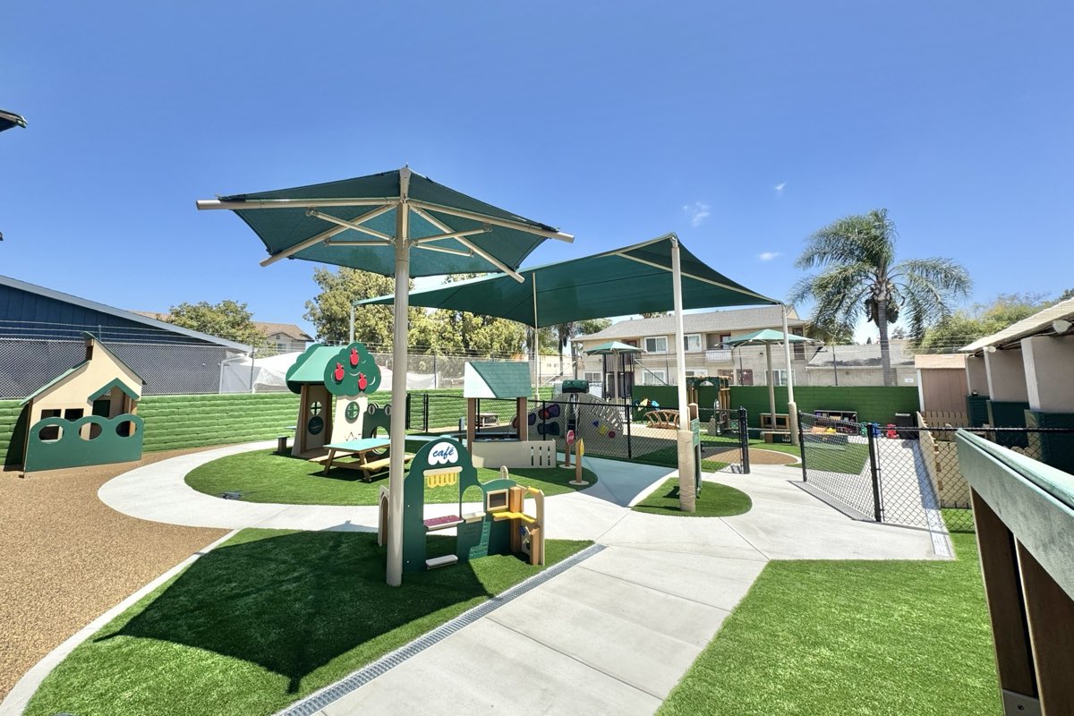 Village Play themed mini-town — small pitched-roof buildings with a cafe service panel, under shade umbrellas